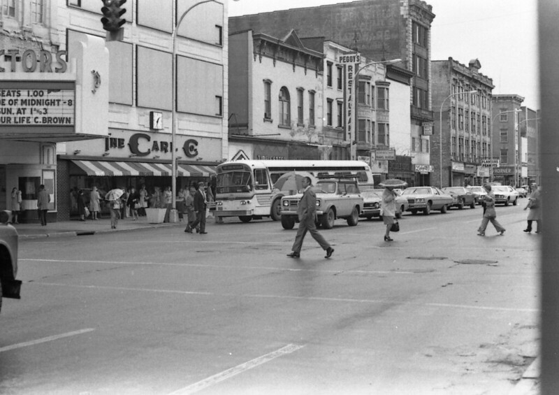 Photograph of a downtown streetscape in Schenectady, New York, 1979. A bit of a theater marquee is seen at the left of the picture, then a row of old downtown buildings in a jumble of sizes and styles. Cars and a city bus are stopped at a red light while pedestrians, some carrying umbrellas, cross the street. On the left side is a sign featuring the logo of The Carl Company, in its headquarters building.