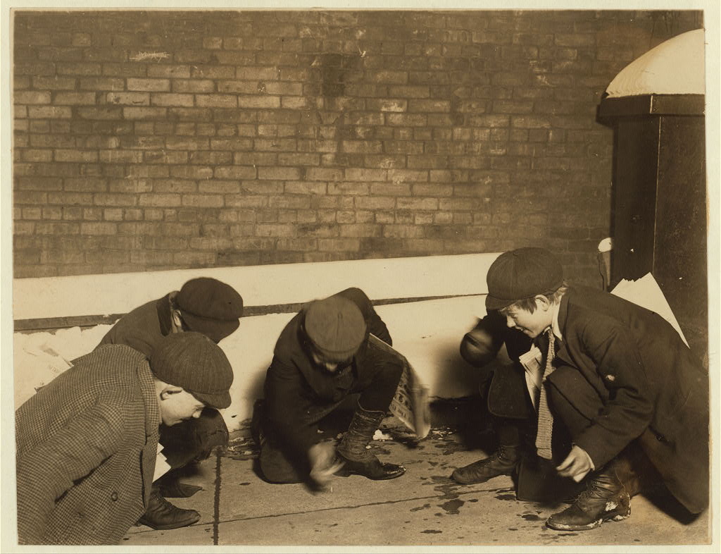 The same newsboys from the other Hine photograph, playing a dice game called craps. They are squatting or kneeling in a semi-circle on a sidewalk, surrounded by snow. 