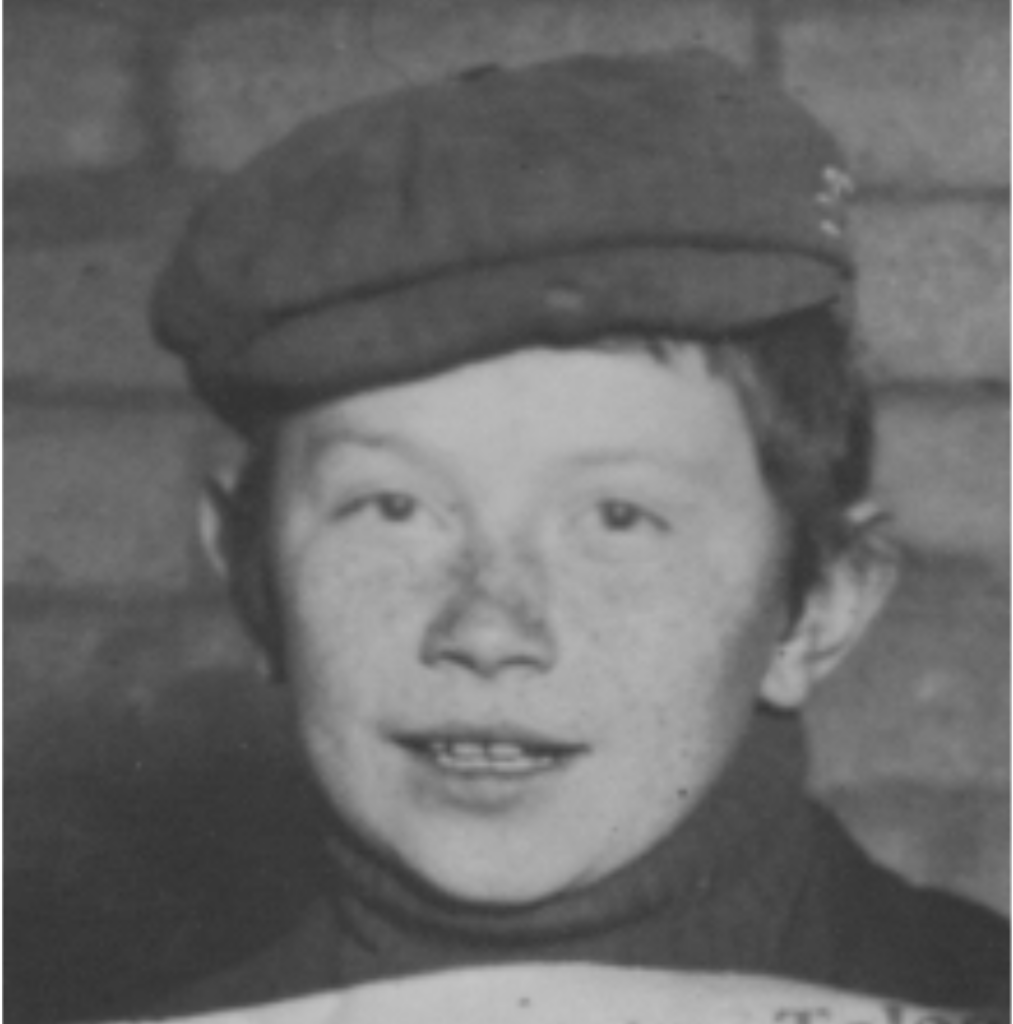 A freckle-faced boy in a cloth cap, hair cut short, facing the camera with a slight smile.