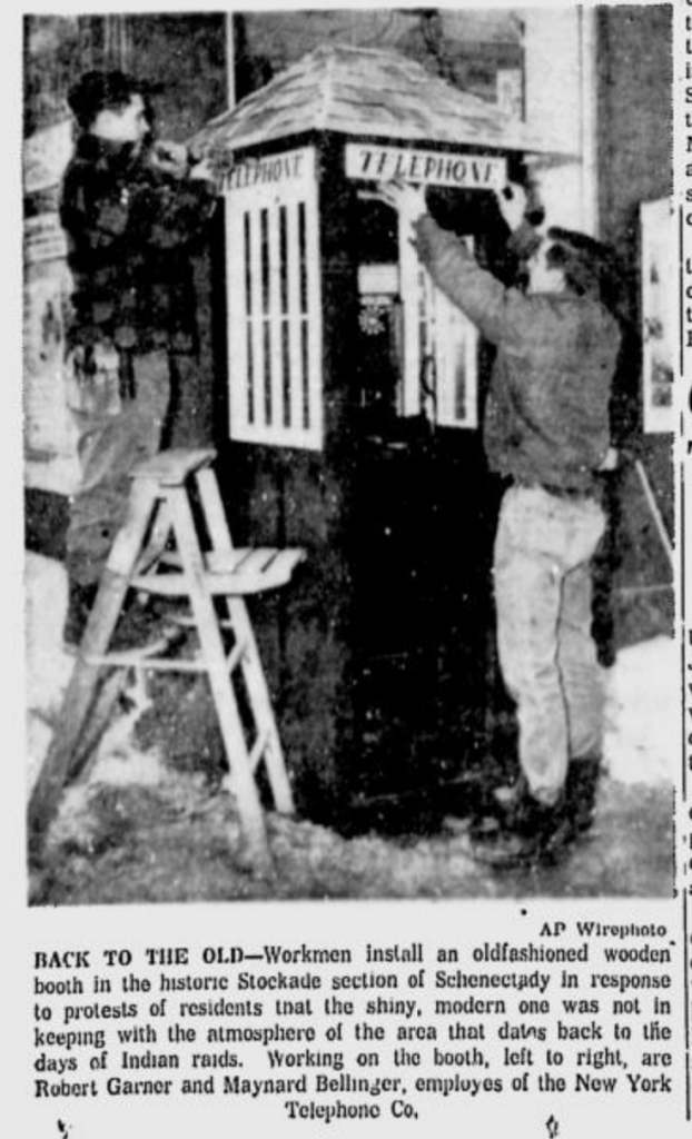 Photograph from a newspaper showing two workers putting finishing touches on a small wooden enclosure, with a small shingled roof, for a telephone booth in Schenectady's Stockade area. One is standing on a short step-latter toward the left wall of the booth and putting up a sign that says "telephone," the other is standing before the booth's door and holding up an identical sign in the spot where it is to be placed. The caption reads "Back to the old – Workmen install an oldfashioned wooden booth in the historic Stockade section of Schenectady in response to protests of residents that the shiny, modern one was not in keeping with the atmosphere of the area that dates back to the days of Indian raids. Working on the booth, left to right, are Robert Garner and Maynard Bellinger, employes of the New York Telephone Co."